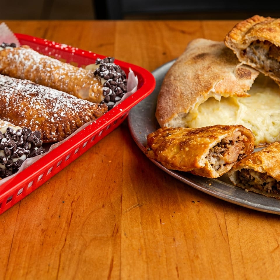 A red basket of cannolis and a metal tray of calzones and empanadas on a wooden table.