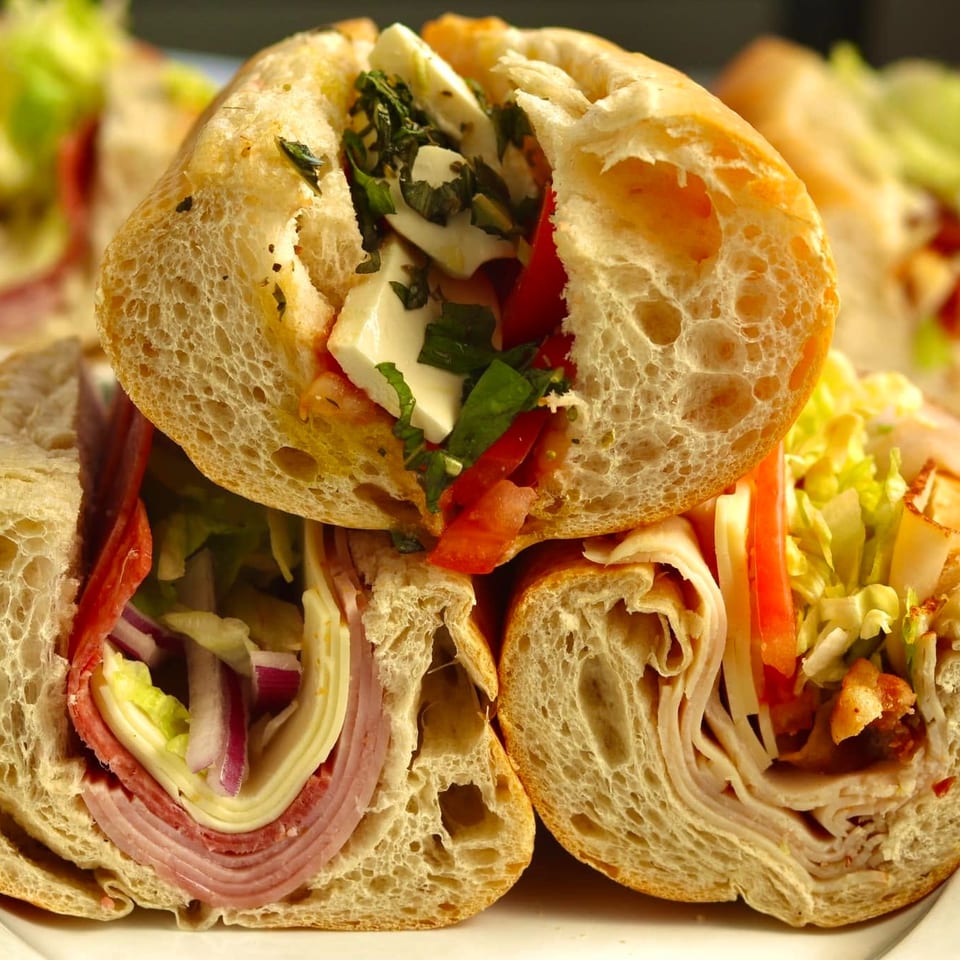 Assorted Italian submarine sandwiches on a plate, filled with deli meats, cheese, lettuce, tomatoes, peppers, and herbs.