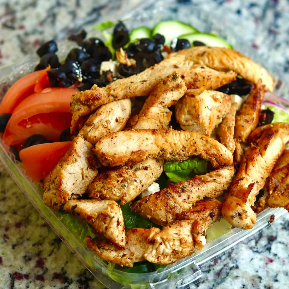 Grilled seasoned chicken strips fill a salad container with lettuce, tomatoes, black olives, cucumber, and red onion.