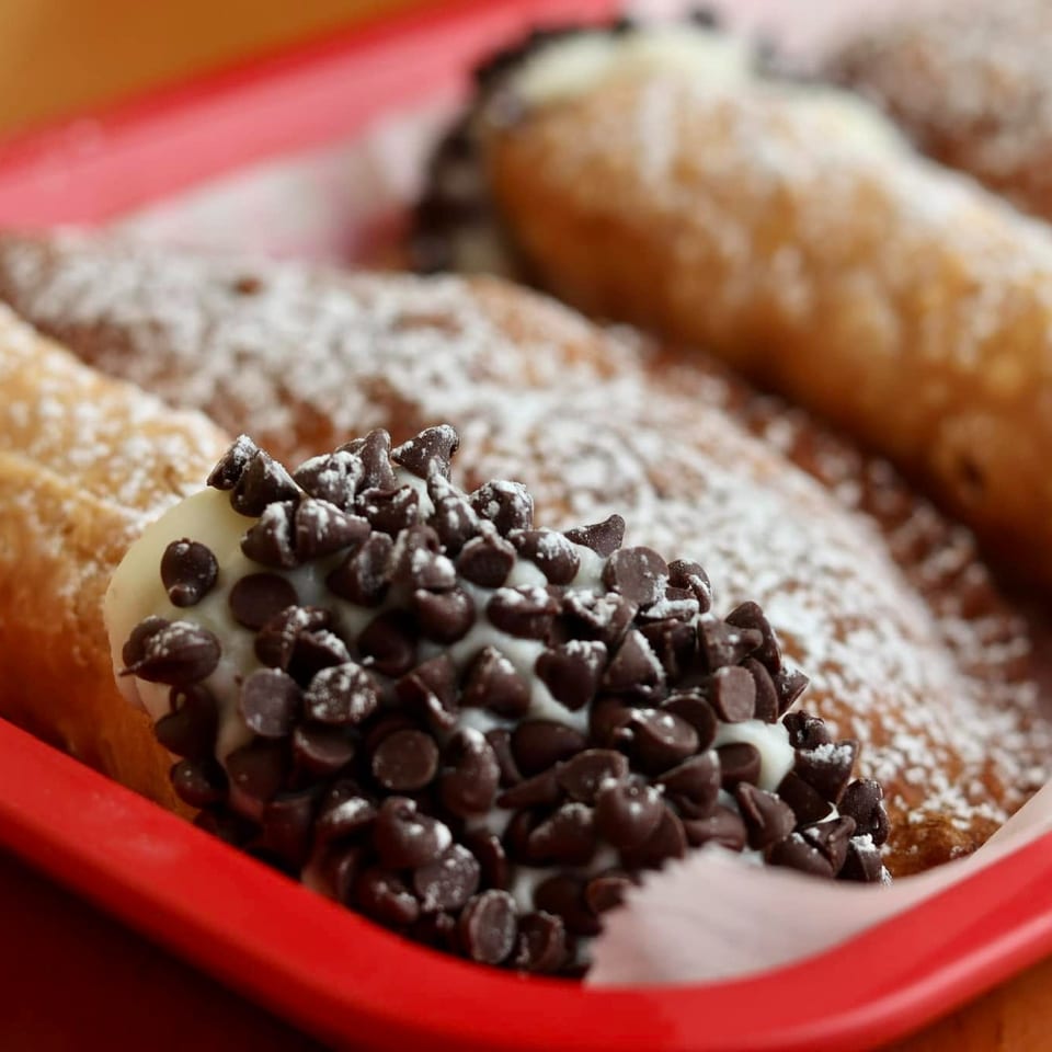 Cannoli pastries dusted with powdered sugar, including one filled with cream and covered in chocolate chips, arranged in a red tray.