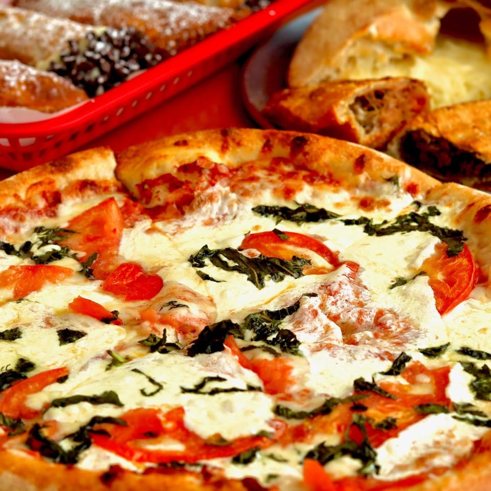 Fresh Margherita pizza topped with melted mozzarella, tomatoes, and basil, surrounded by assorted pastries and baked goods.